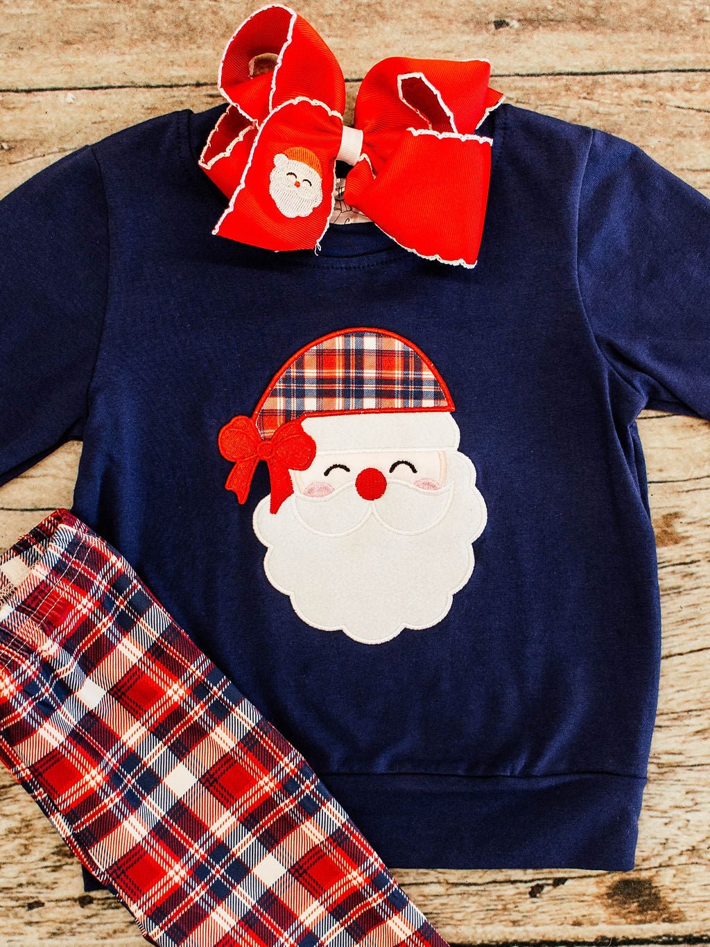 close up of a navy long sleeve with a santa wearing a plaid hat applique on the center and red, navy, plaid pants showing in corner, styled with red bow featuring a santa embroidery on it