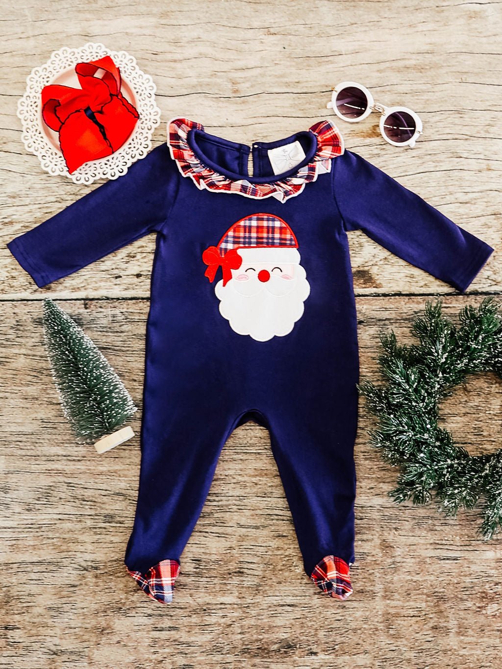 flay lay of a navy long sleeve romper with a santa wearing a plaid hat applique in center, red navy and white plaid ruffled collar and footies