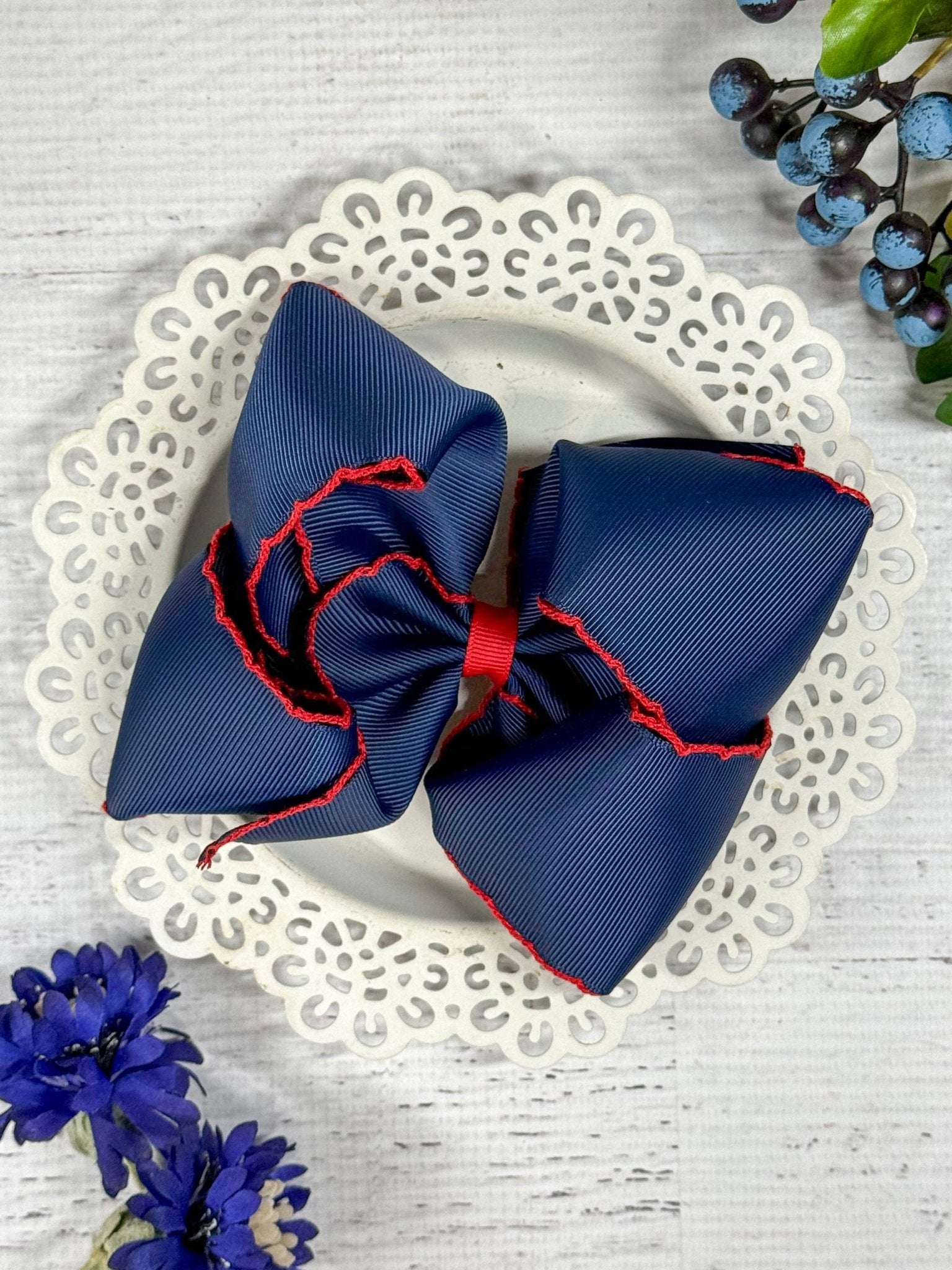 A navy blue hair bow with red moonstitch detailing on a white lace plate, surrounded by blue and purple flowers.