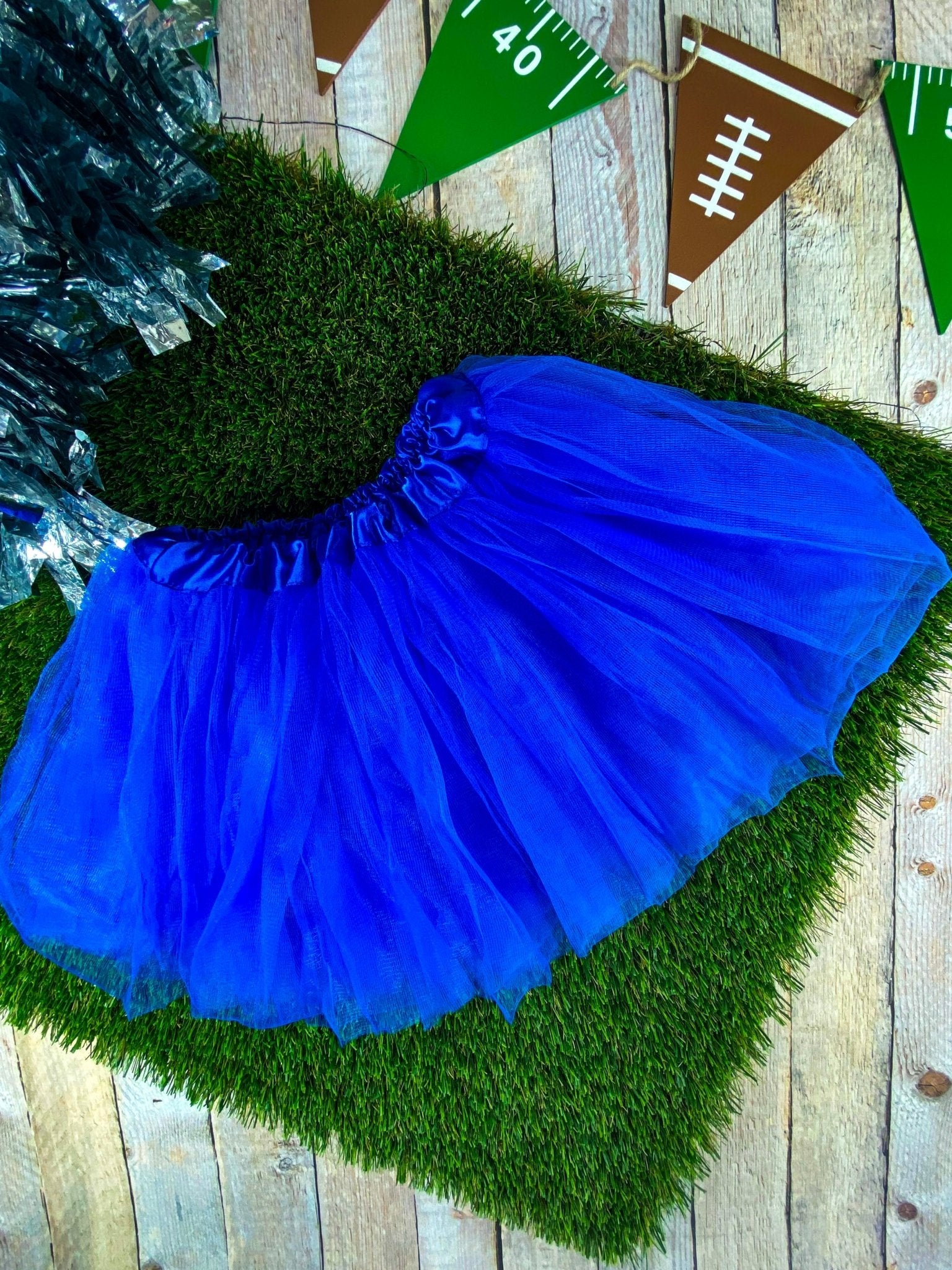 A blue baby tutu skirt laid out on a wooden surface with artificial grass, decorations in the background.