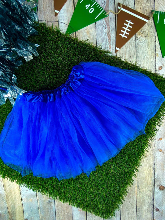 A blue baby tutu skirt laid out on a wooden surface with artificial grass, decorations in the background.