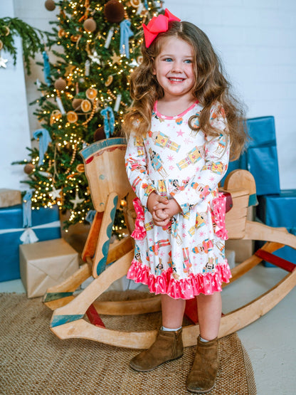 Model wearing the Nutcracker Galore Twirl Dress with Pockets shown worn in a festive holiday setting. The long-sleeve dress has a white background with bright nutcracker designs, pink star-trimmed ruffles, and soft gathered fabric perfect for twirling. Styled with a large bow and brown ankle boots for a cozy Christmas look.