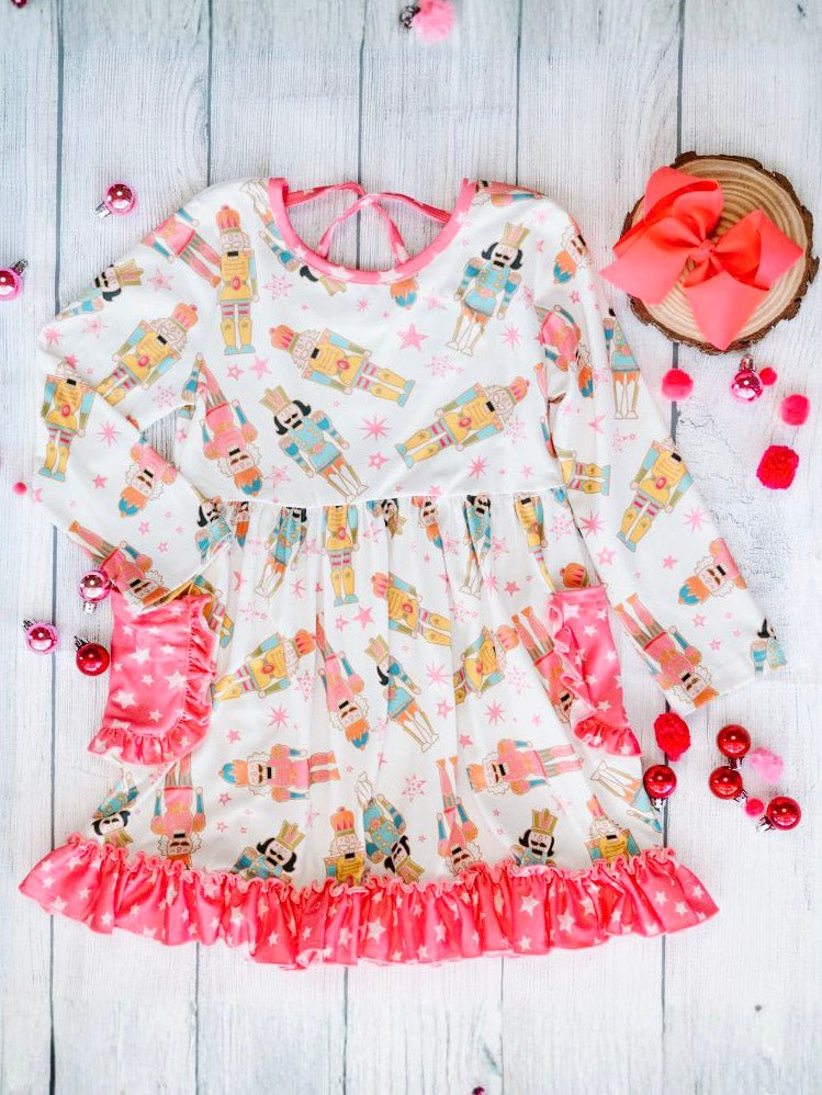 Full flat lay of the Nutcracker Galore Twirl Dress with Pockets displayed on a light wood background. The dress features colorful nutcracker illustrations, long sleeves, pink star-print pockets, and a matching ruffled hem for a playful holiday design.