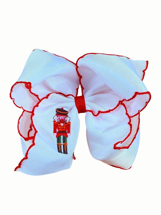 White moonstitch hair bow with red stitched edges and an embroidered nutcracker soldier on the tail, on a white background