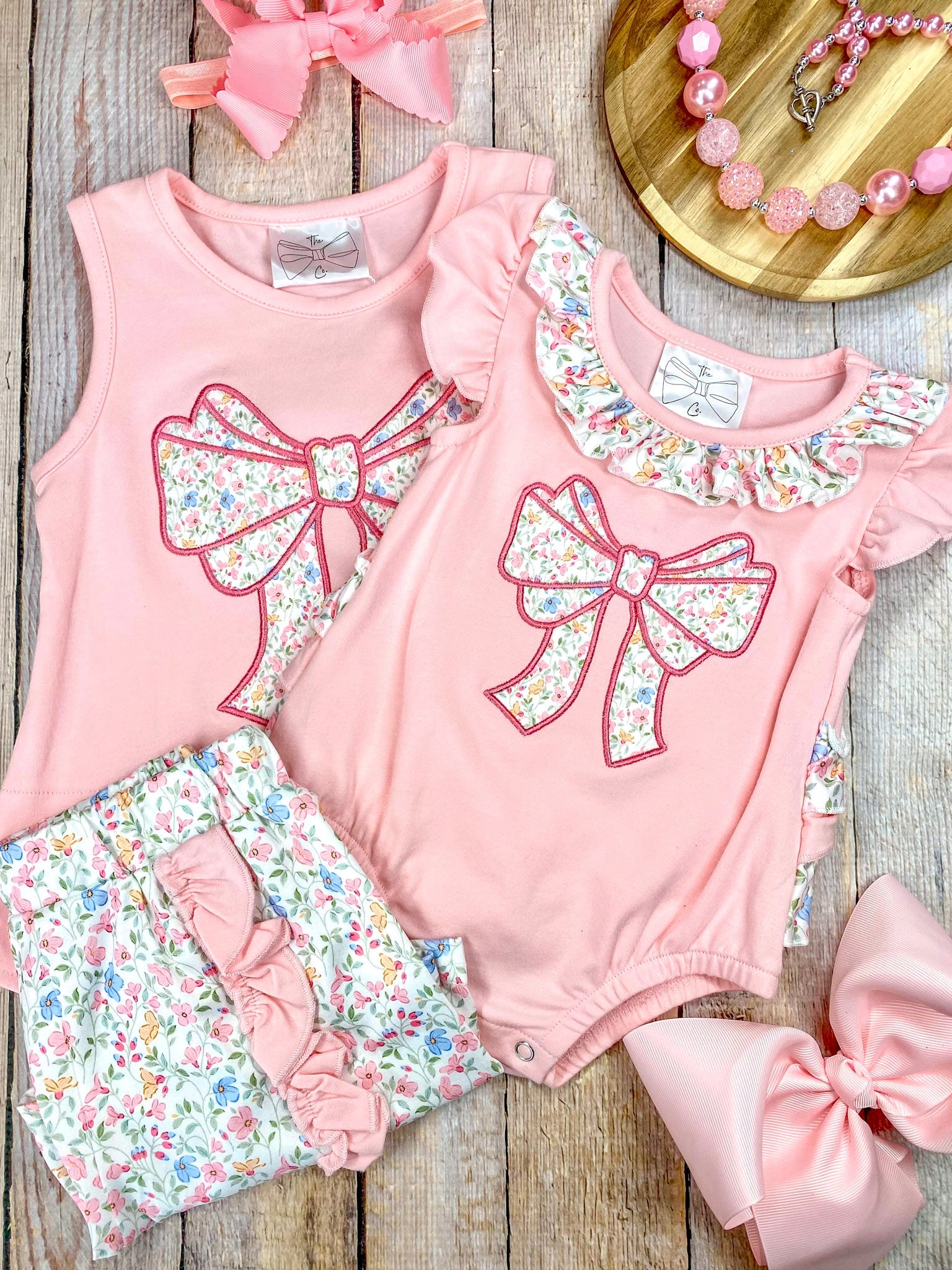 Pink sleeveless top with a large bow and matching floral shorts and infant romper on wooden background