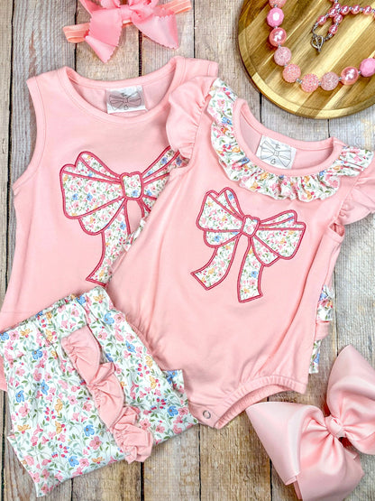 Pink sleeveless top with a large bow and matching floral shorts and infant romper on wooden background