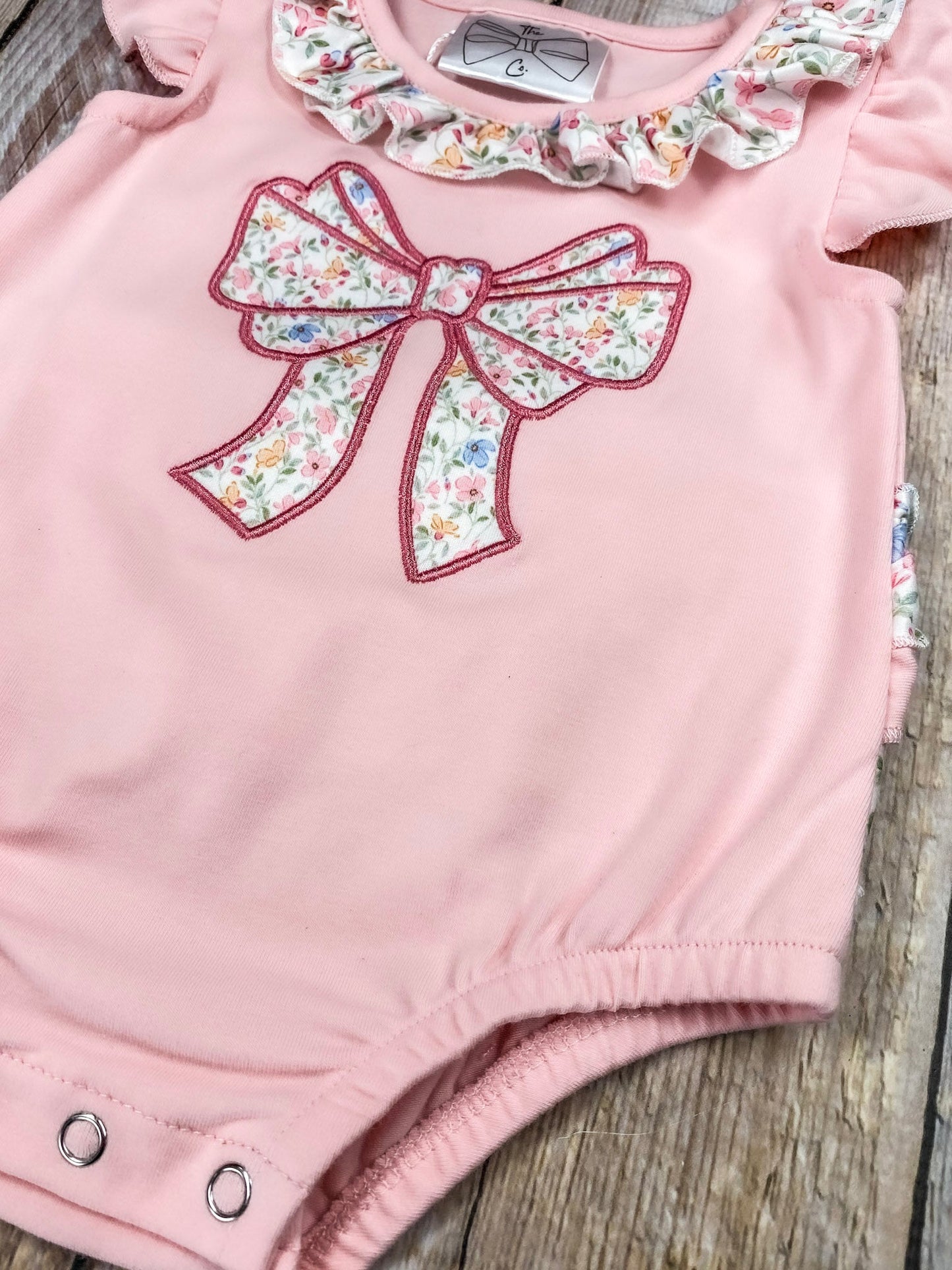 Pink baby romper with floral bow and ruffles on a wooden background