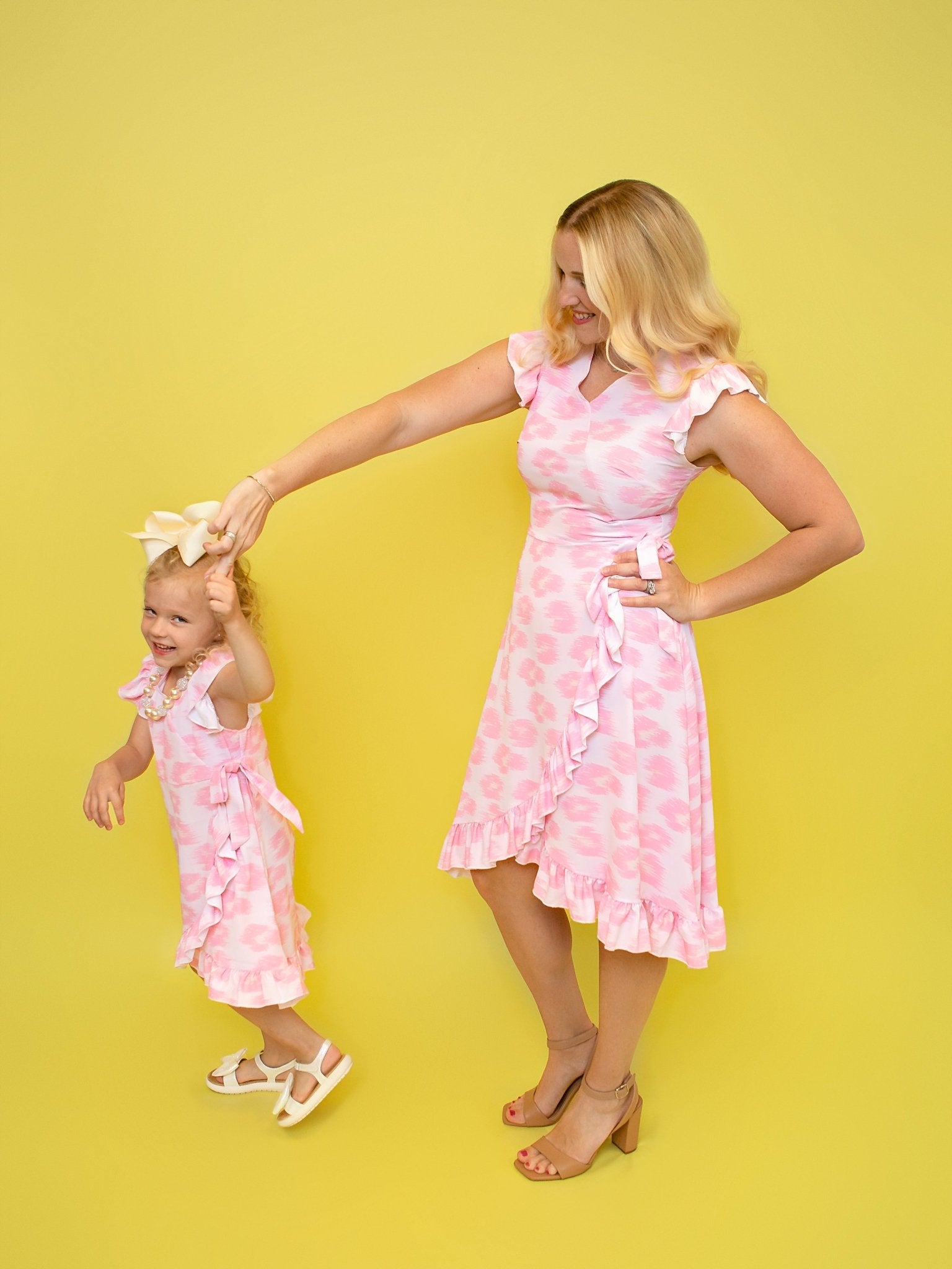  mother and daughter, wearing matching pink ruffled dresses with floral patterns, standing against a yellow background.