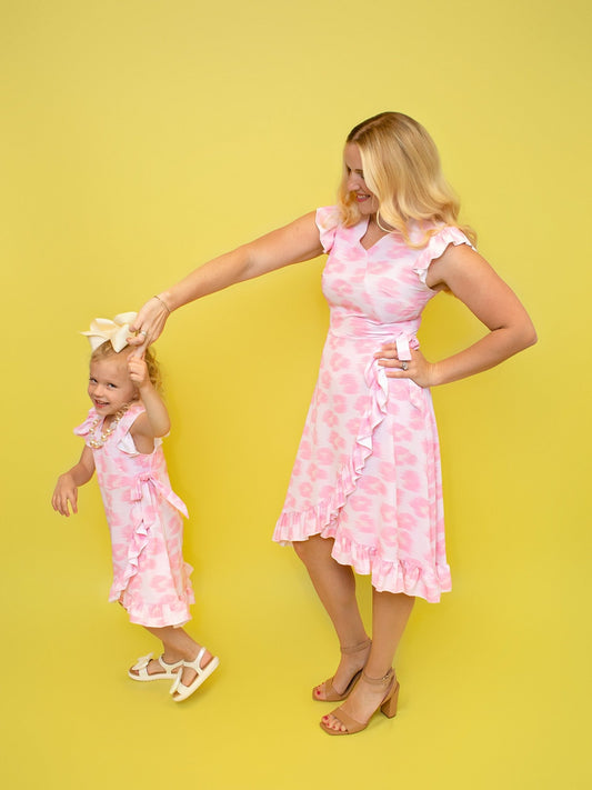  mother and daughter, wearing matching pink ruffled dresses with floral patterns, standing against a yellow background. - Alternative view