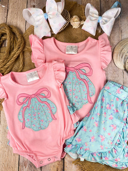 Pink baby romper and matching shorts set with a blue bow design on a wooden background