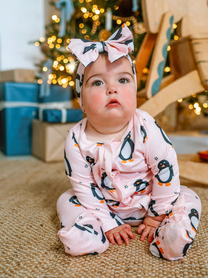 Baby in a pink penguin onesie with a headband, sitting on a carpeted floor with Christmas decorations in the background.
