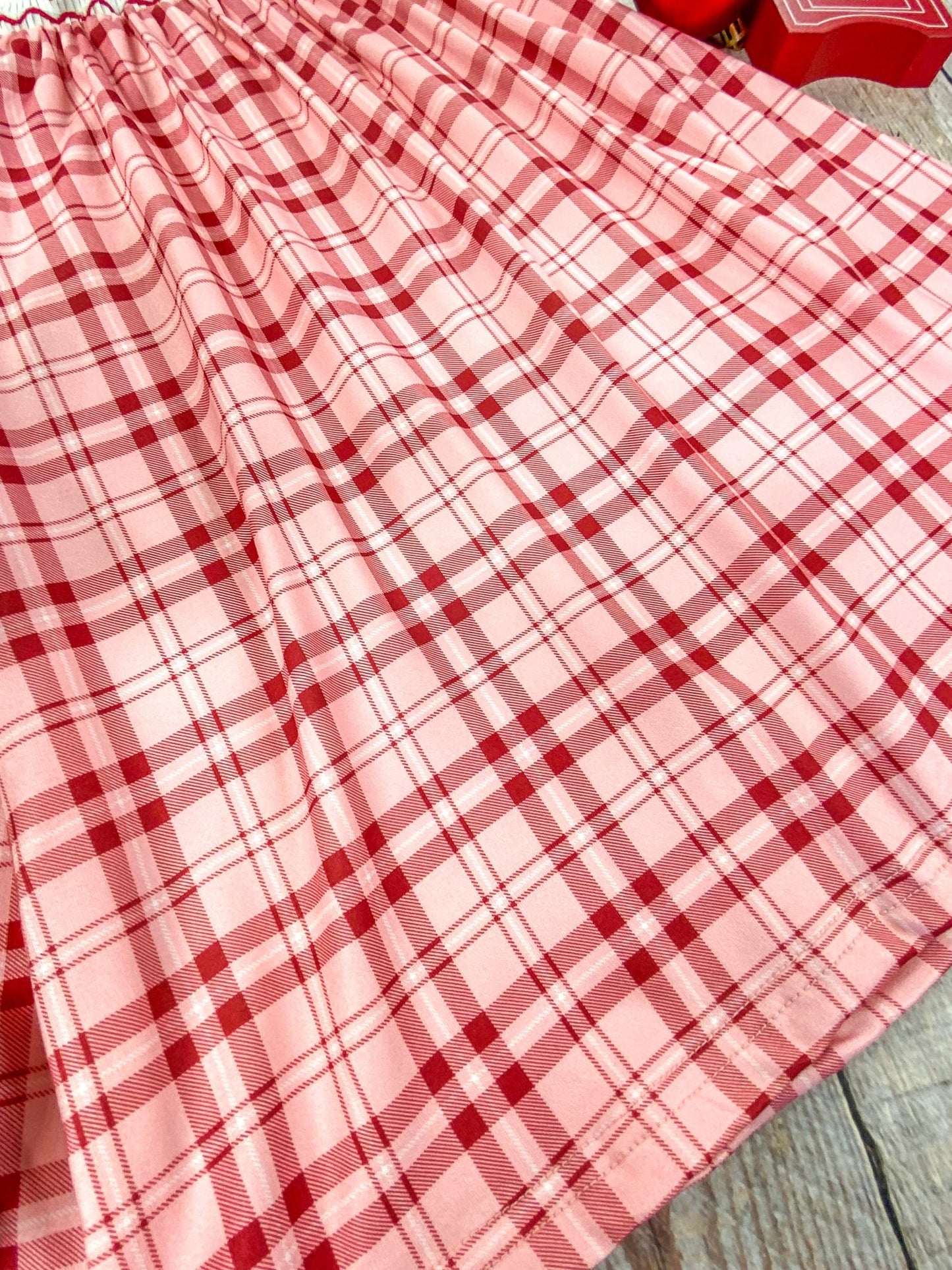 Close-up of the bottom hem of a red and pink plaid Christmas dress. The soft pleats and lightweight fabric are shown in detail, highlighting the gentle flow and texture of the dress against a wooden background.