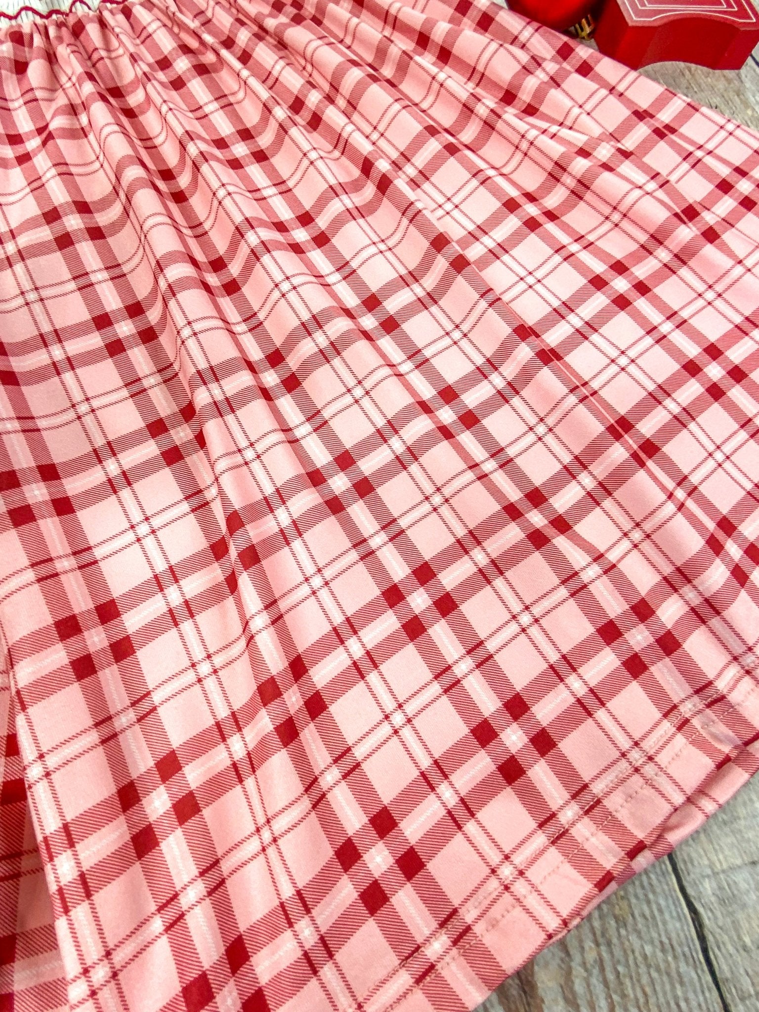 Close-up of the bottom hem of a red and pink plaid Christmas dress. The soft pleats and lightweight fabric are shown in detail, highlighting the gentle flow and texture of the dress against a wooden background.