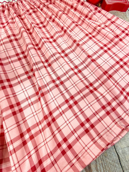 Close-up of the bottom hem of a red and pink plaid Christmas dress. The soft pleats and lightweight fabric are shown in detail, highlighting the gentle flow and texture of the dress against a wooden background.