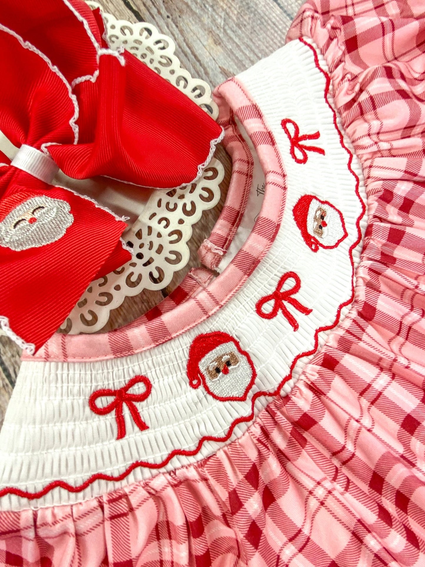 Close-up of a white smocked collar on a red and pink plaid Christmas dress, embroidered with two Santa Claus faces and 3 red bows. The detailed stitching are clearly visible, emphasizing the festive and delicate craftsmanship.