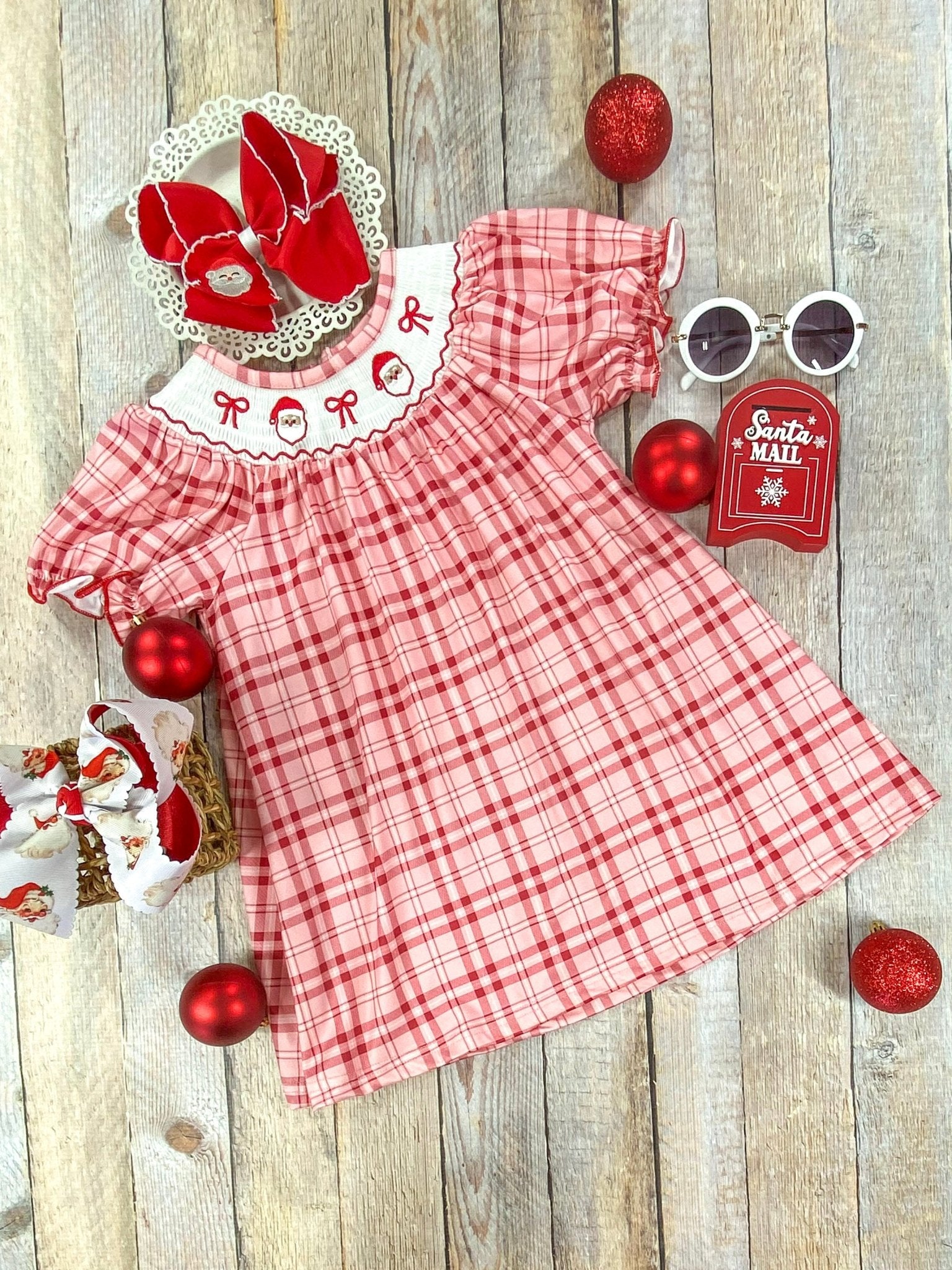 Red and pink plaid girl's Christmas dress with puff sleeves and a white smocked collar embroidered with three Santa faces and red bows. A large red bow accents one sleeve. The dress is laid flat on a wooden background, styled with festive red ornaments.