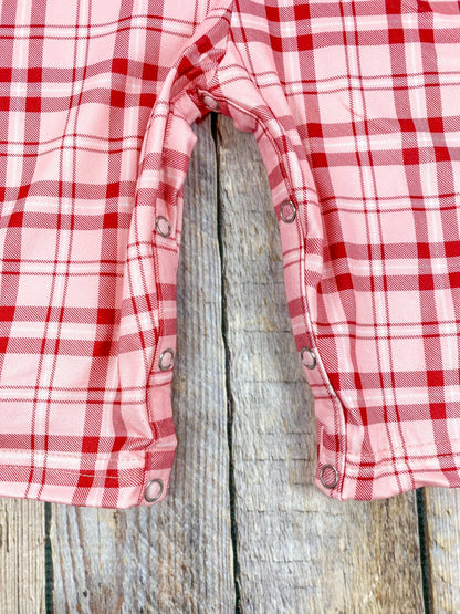 Close-up of the legs of a red and pink plaid baby boy’s romper, showing snap closures along the inner seams for easy diaper changes. The fabric’s soft texture and clean stitching lines are clearly visible, laid flat on a wooden background.