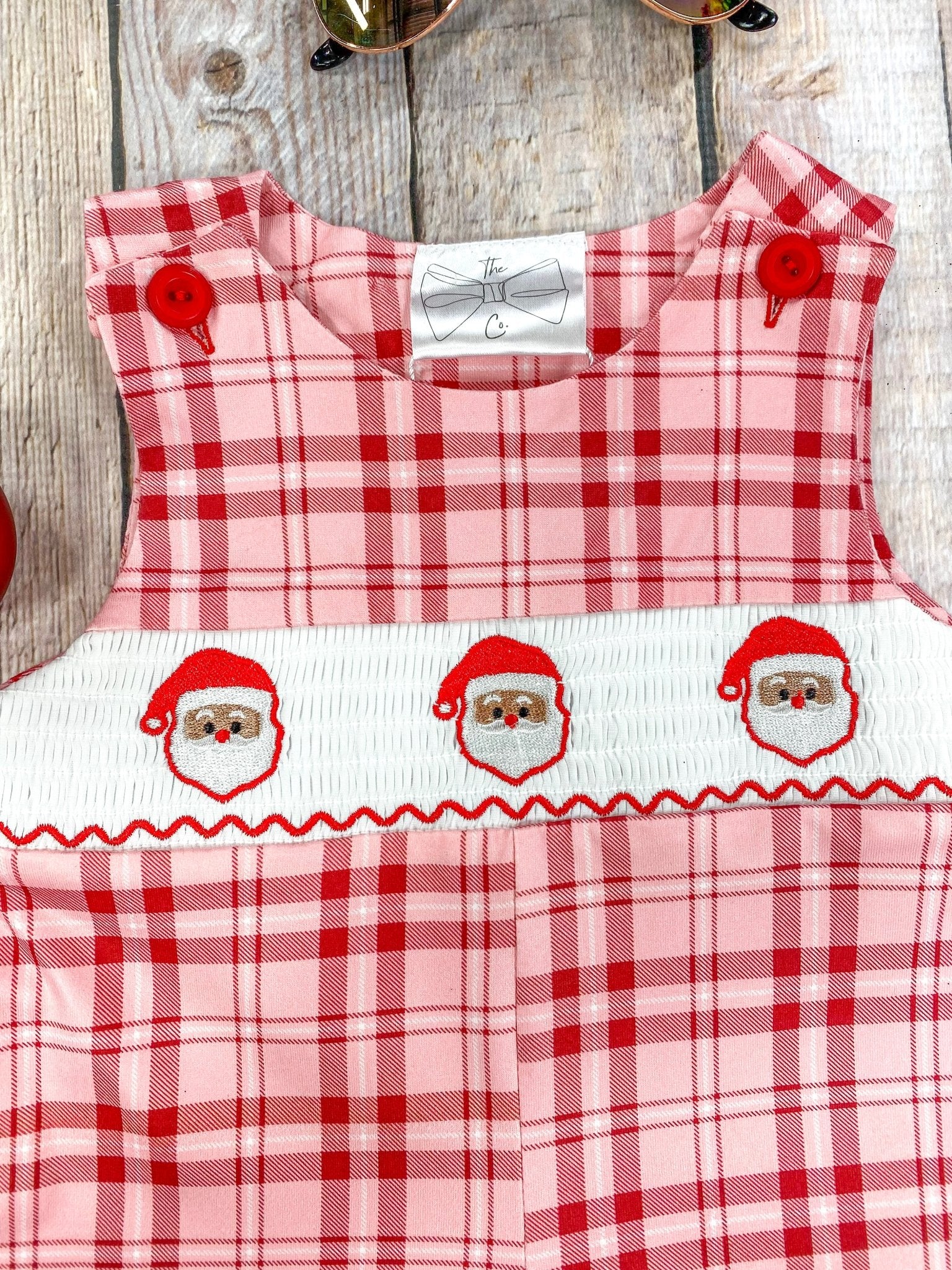 Close-up of the top portion of a baby boy’s red and pink plaid romper with a white smocked panel featuring three embroidered Santa Claus faces. Red buttons secure the shoulder straps, and fine stitching details are visible on the plaid fabric.
