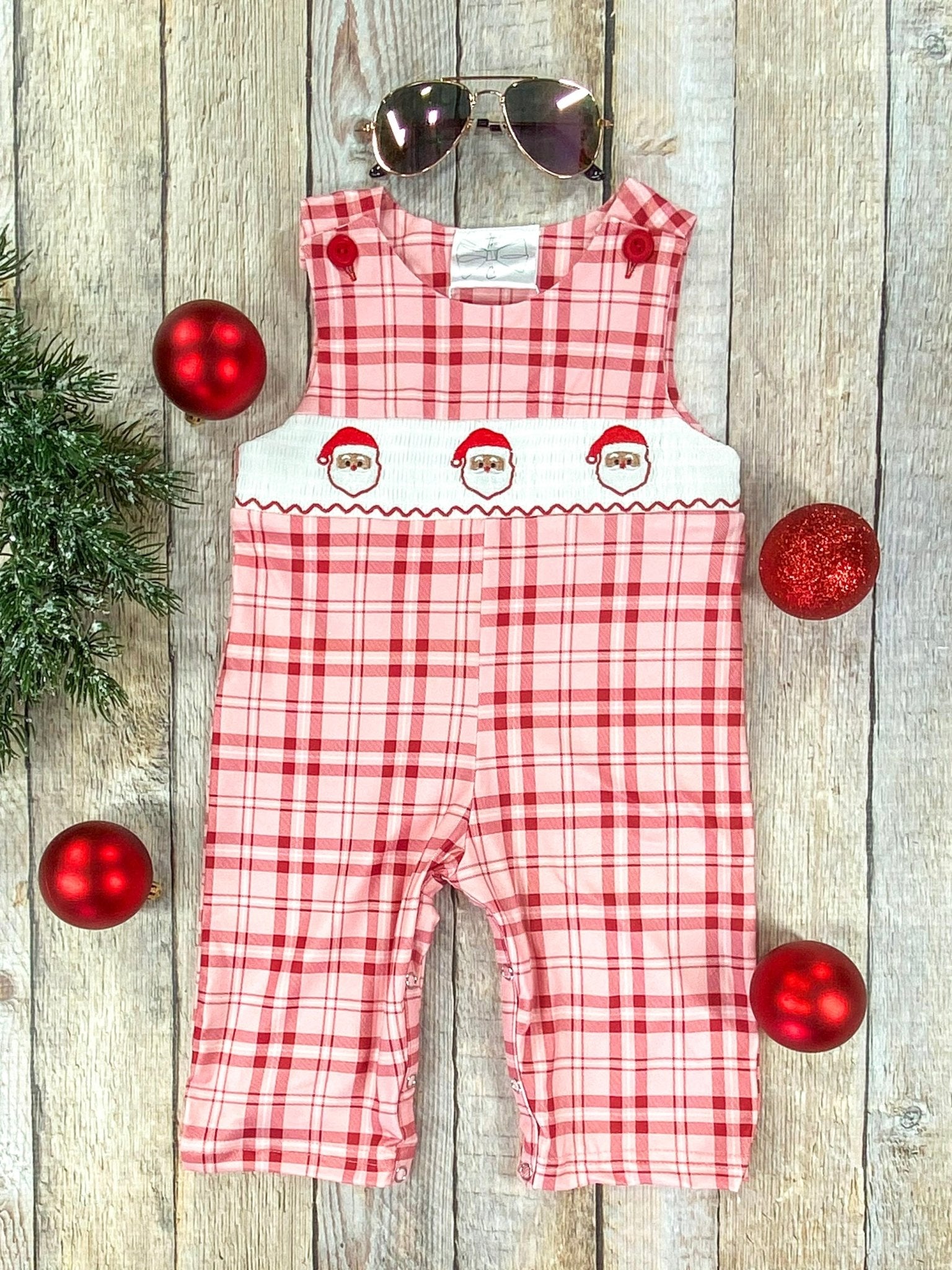 Flat lay of a red and pink plaid long romper for a baby boy with a smocked white chest panel featuring 3embroidered Santa faces. The outfit has red buttons on the straps and is styled with aviator sunglasses, red ornaments, and greenery on a wooden background.