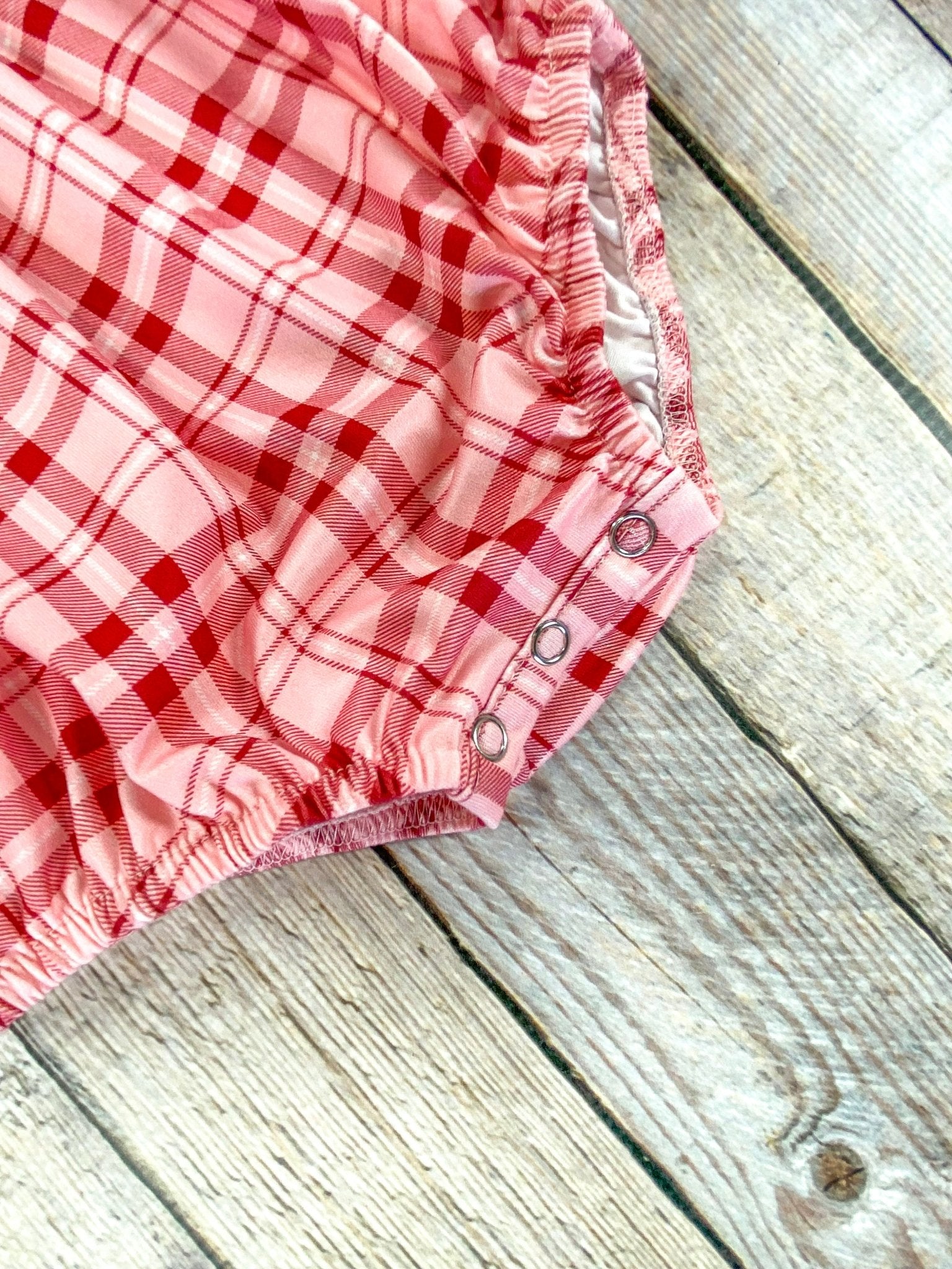 Close-up of the bottom of a red and pink plaid infant romper showing two metal snaps along the leg opening. The elastic leg cuffs and soft fabric texture are clearly visible, highlighting ease of diaper changes.