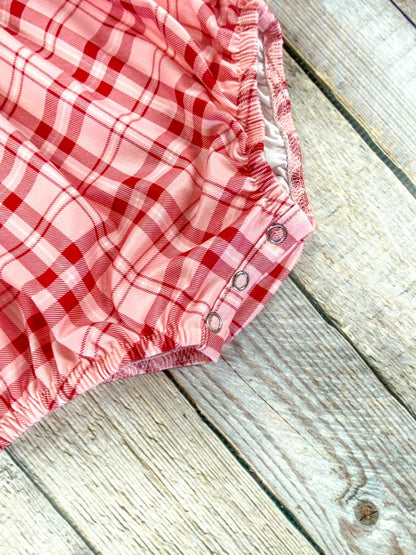 Close-up of the bottom of a red and pink plaid infant romper showing two metal snaps along the leg opening. The elastic leg cuffs and soft fabric texture are clearly visible, highlighting ease of diaper changes.