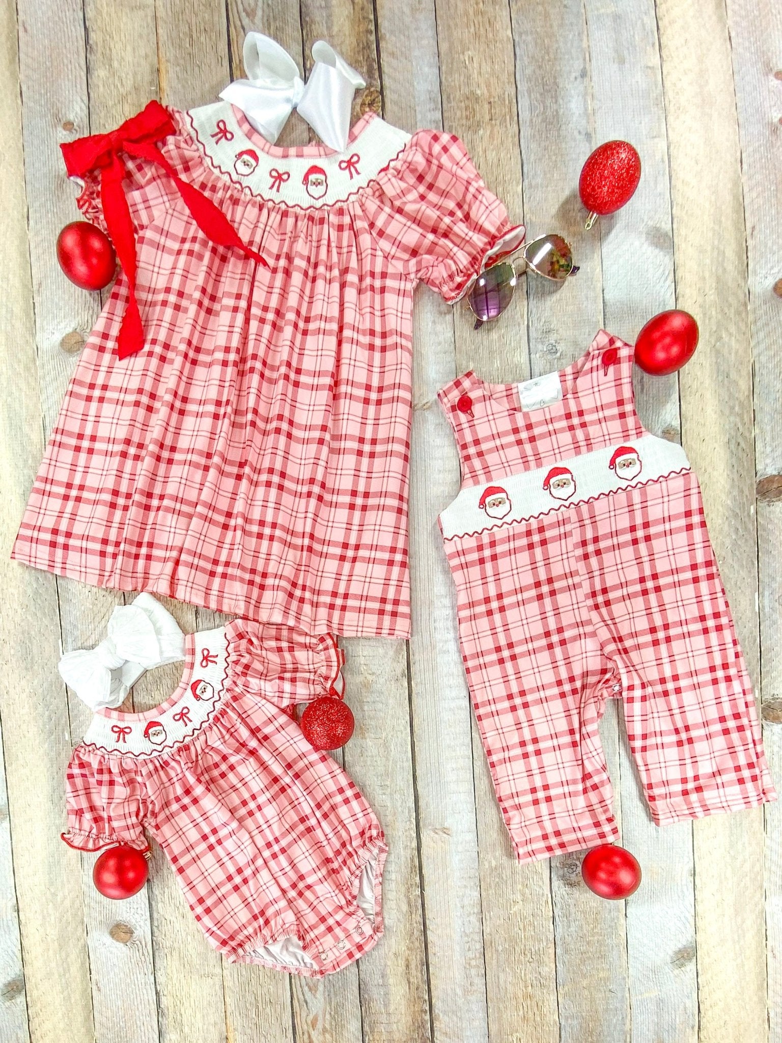Flat lay of three coordinating children's Christmas outfits on a wood plank background. Outfits are made from red and pink plaid fabric with white collars embroidered with Santa faces and red bows. Includes a short-sleeve dress with a red bow, a romper with puff sleeves, and a long romper overall with red buttons. Surrounded by red Christmas ornaments and festive accessories.