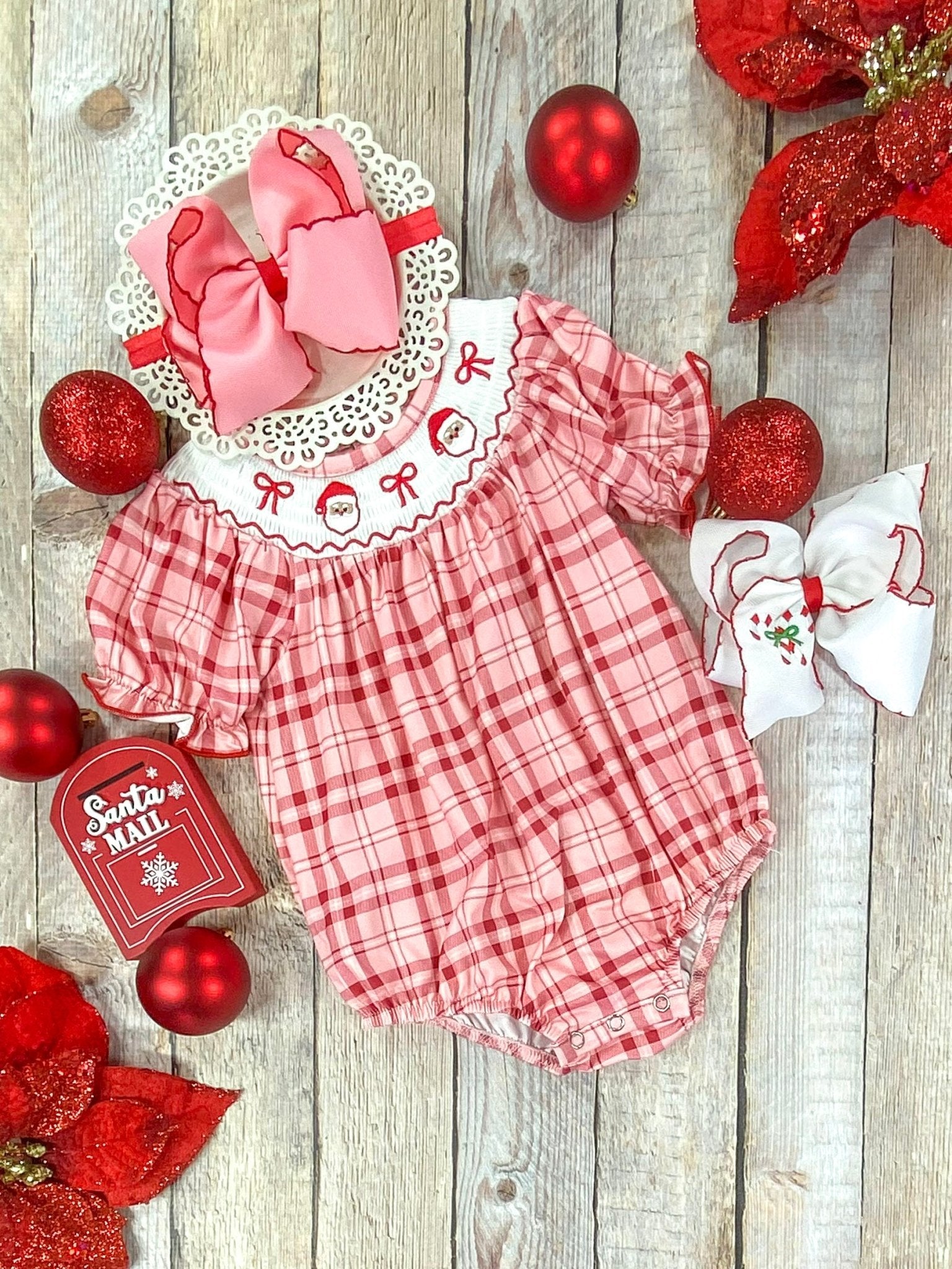 A close-up flat lay of a baby girl's Christmas romper in red and pink plaid with puff sleeves and a white smocked collar featuring embroidered Santa faces and red bows. Styled with large pink and white hair bows, red ornaments, poinsettias, and a decorative "Santa Mail" box on a rustic wooden background.