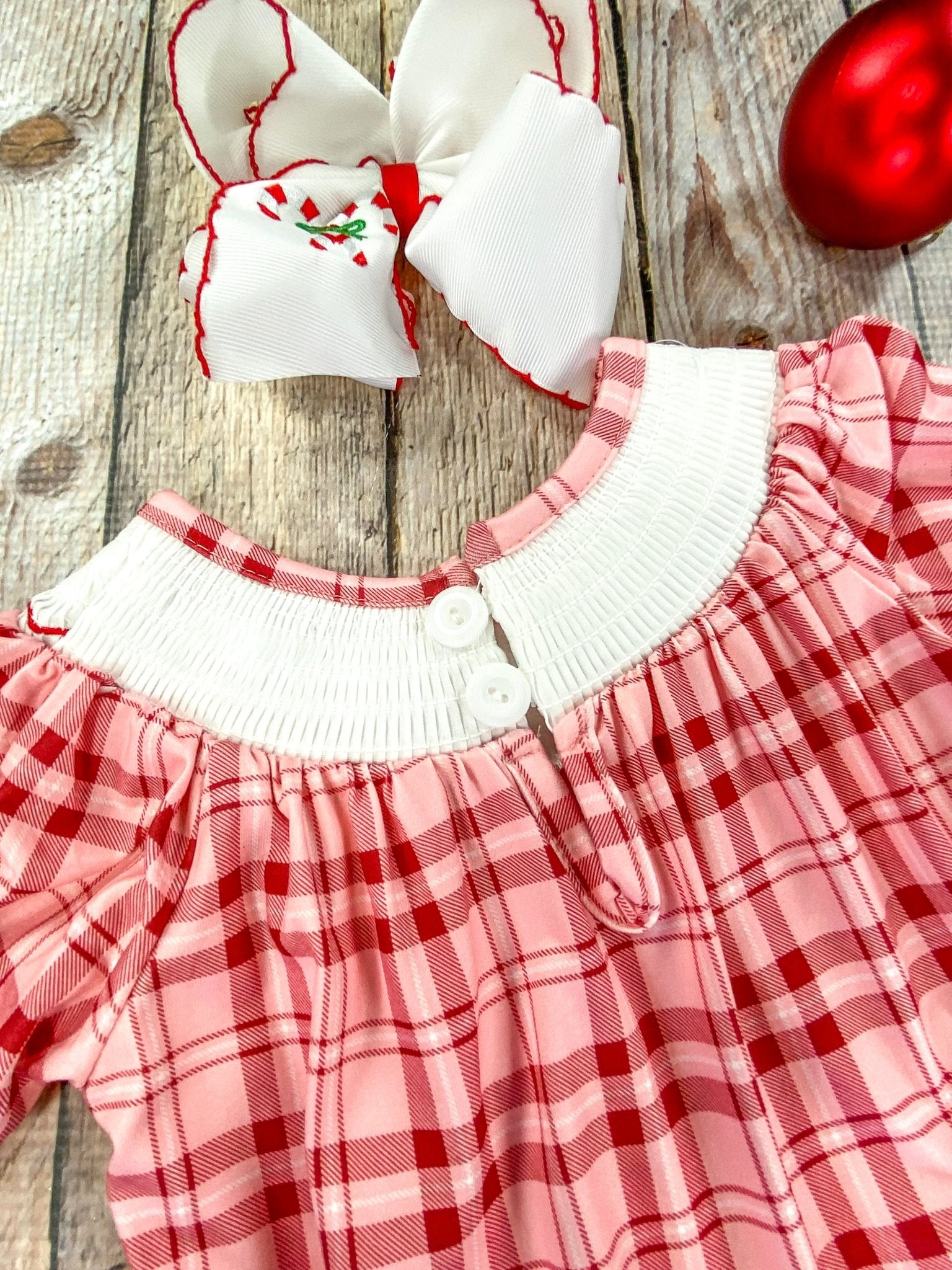 Close-up of the back of a red and pink plaid infant romper featuring a white collar with 2 small buttons for fastening. The gathered fabric and delicate stitching around the neckline are visible, laid flat on a wooden background.