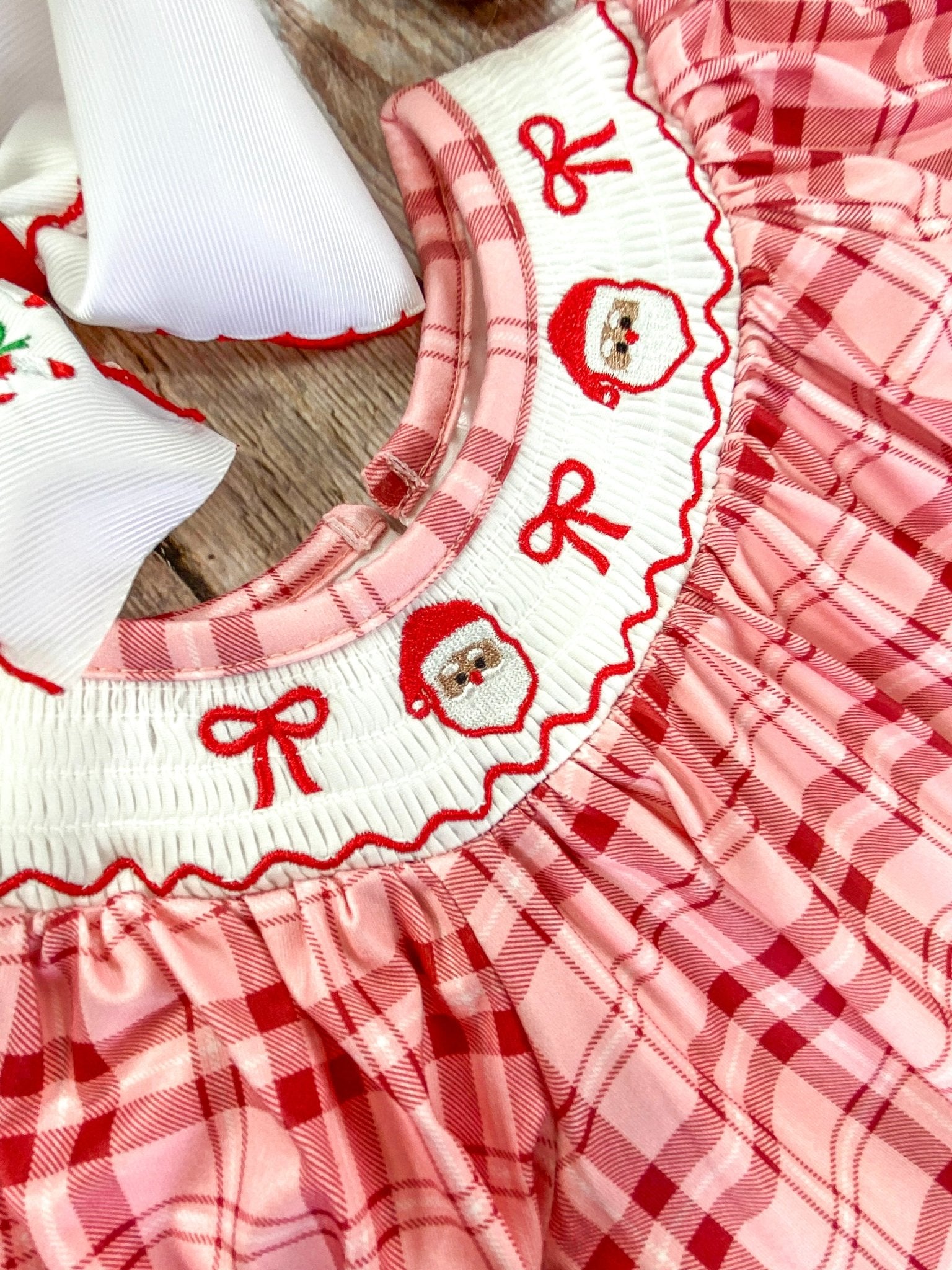 Close-up of a white smocked collar on an infant romper, embroidered with 2 Santa Claus faces and 3 red bows. The detailed stitching emphasizing the festive and delicate craftsmanship.