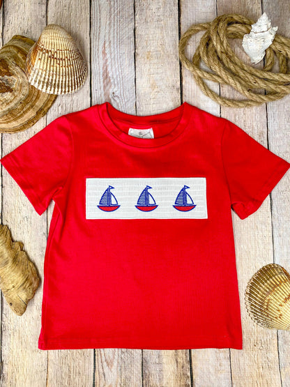 Red t-shirt with a white stripe featuring three blue sailboat graphics on a wooden background