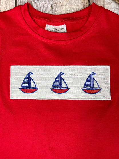 Red t-shirt with a white stripe featuring three blue sailboat graphics on a wooden background