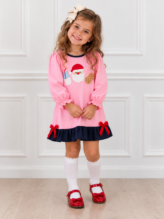 Young girl wearing a pink Santa Christmas dress with milk and cookie appliqués, navy ruffle hem and red bows, paired with white knee-high socks and red Mary Jane shoes.