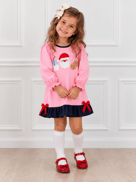Young girl wearing a pink Santa Christmas dress with milk and cookie appliqués, navy ruffle hem and red bows, paired with white knee-high socks and red Mary Jane shoes.