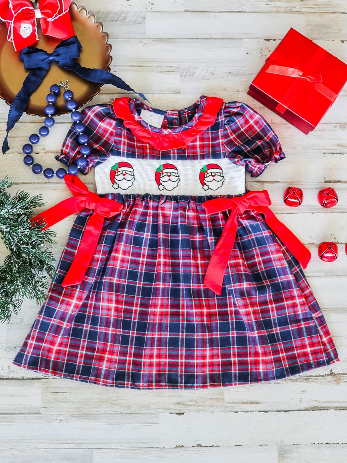 Girls plaid Christmas dress with smocked Santa face, laid flat with holiday decor