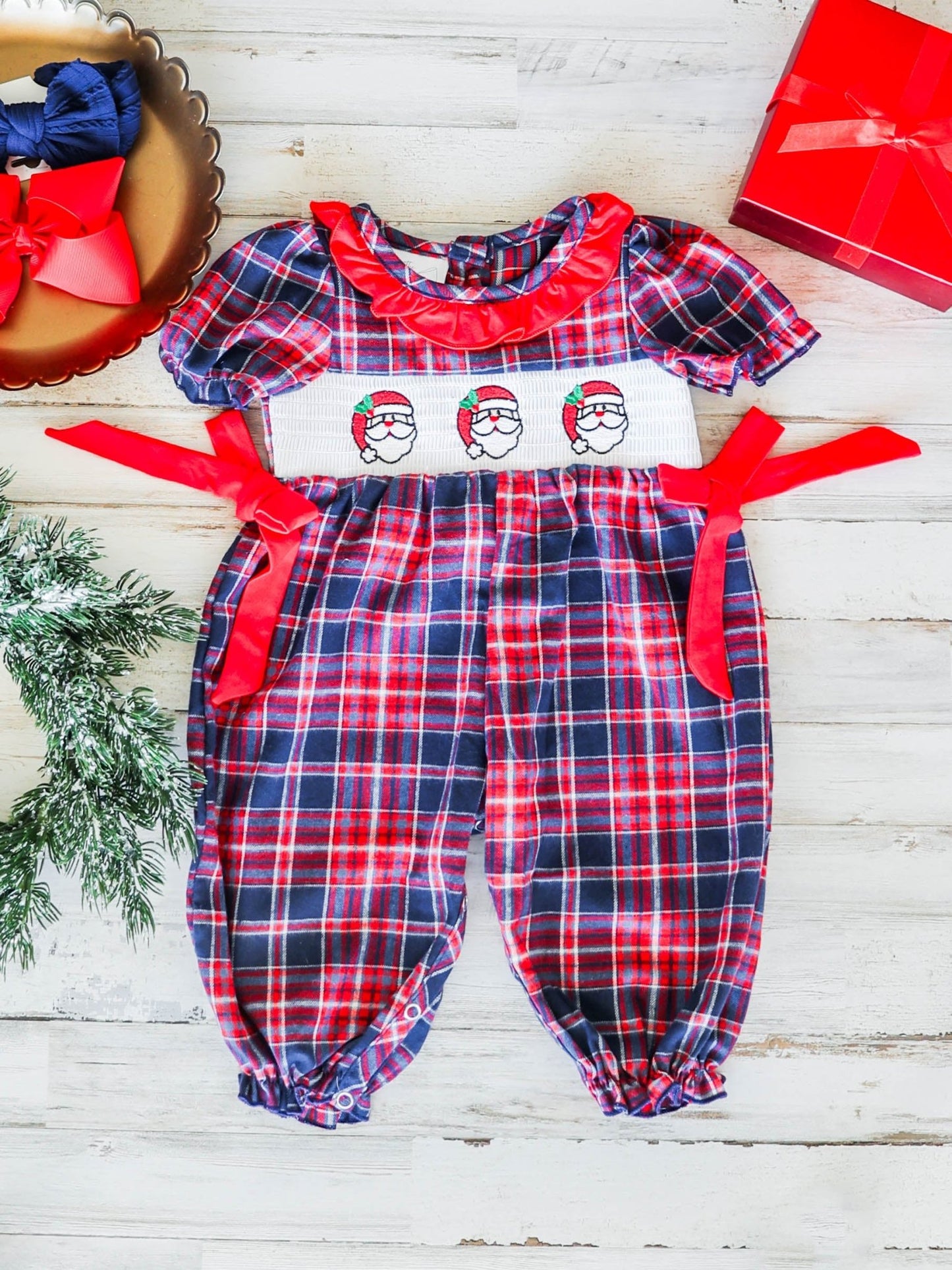 Plaid baby romper with red bows and Santa face design on a wooden surface.