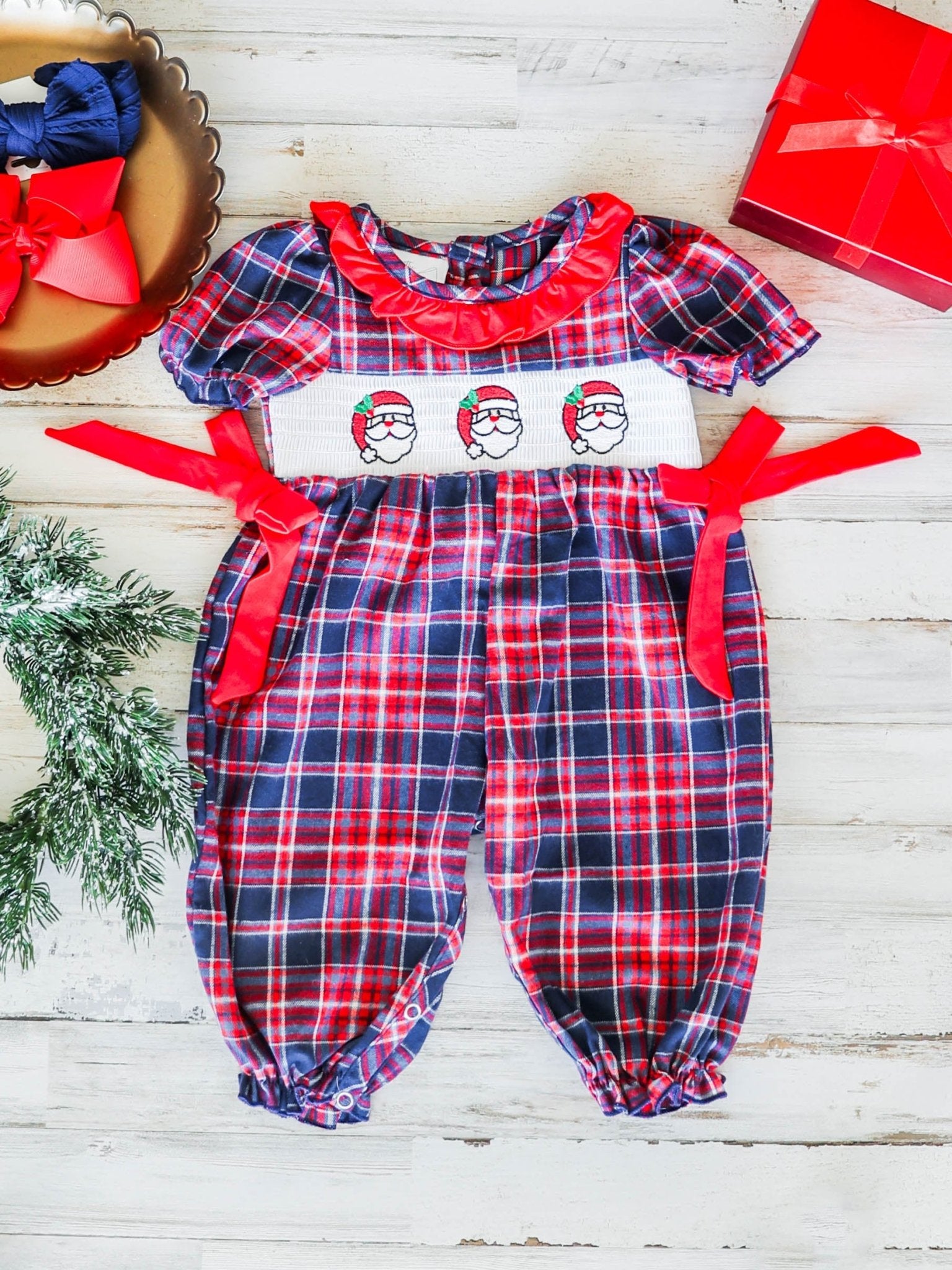 Plaid baby romper with red bows and Santa face design on a wooden surface.