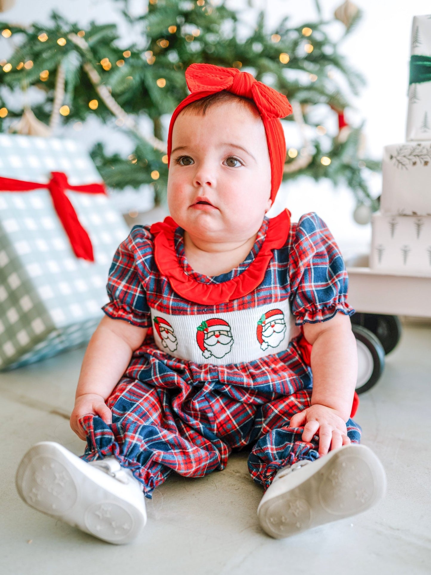 Baby in a festive outfit with Santa Claus design, sitting in front of a Christmas tree.