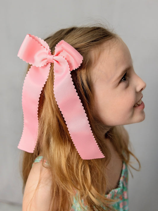 A young girl with long hair wearing a pink scalloped long tail hair bow with an alligator clip.