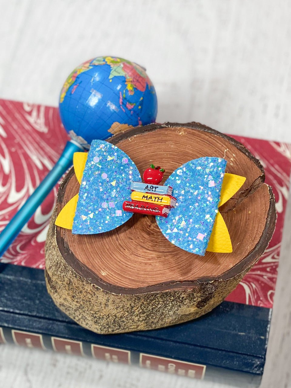 A blue glitter hair bow with a pencil design placed on a wooden slice next to a globe and a pencil.