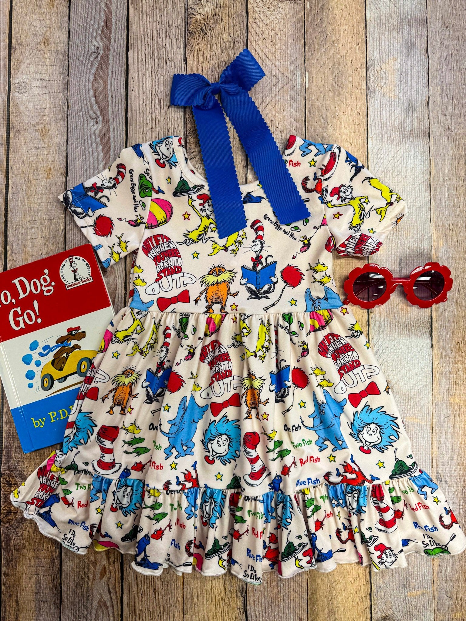 Children's dress with colorful print and blue bow on a wooden surface, accompanied by a book titled 'Dr. Seuss' book'.