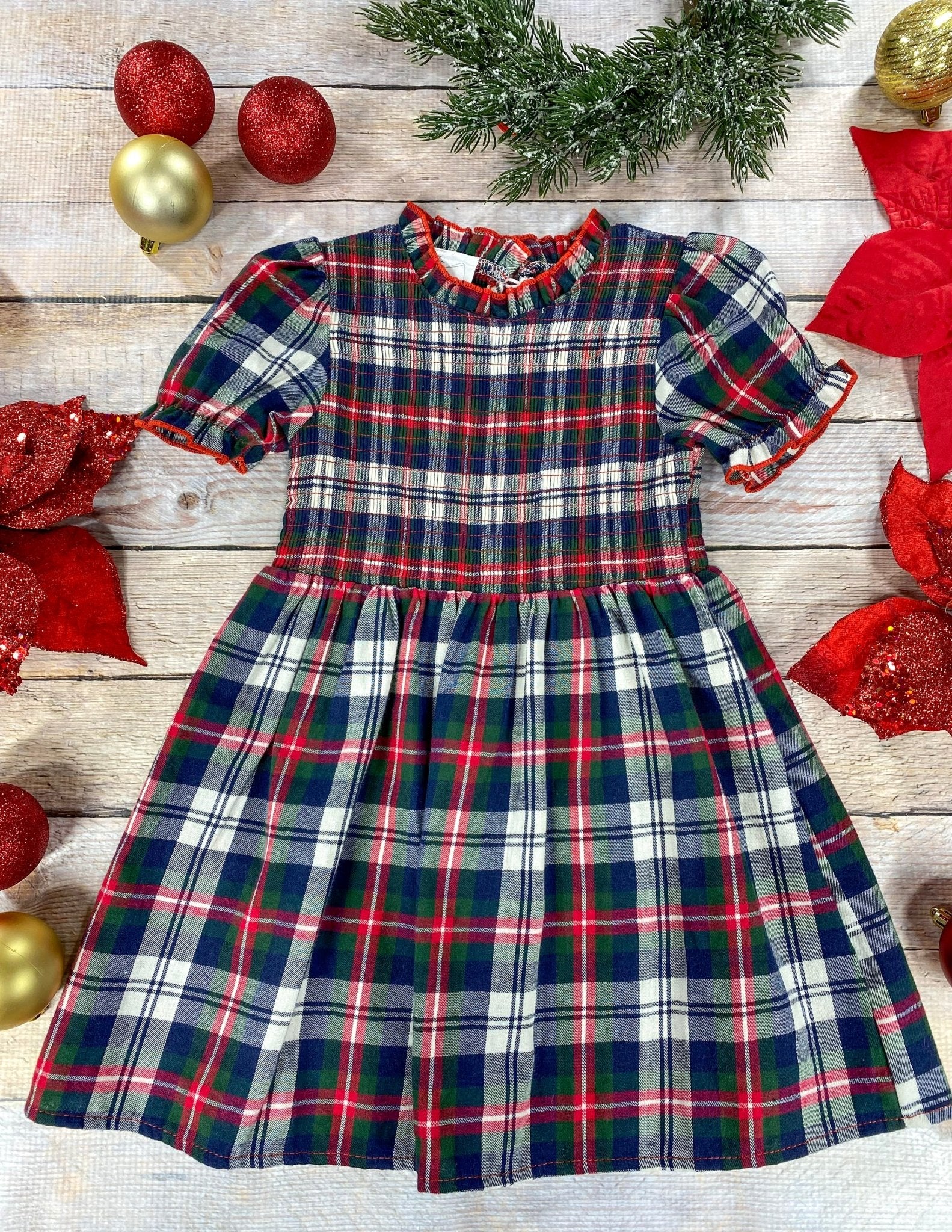 A classic holiday plaid dress for a young girl featuring red, green, navy, and white colors. The dress has puff sleeves, a gathered waist, and a ruffled collar trimmed in red. It’s displayed flat on a wood background surrounded by Christmas decorations, including red poinsettias, glittery red and gold ornaments, and evergreen foliage