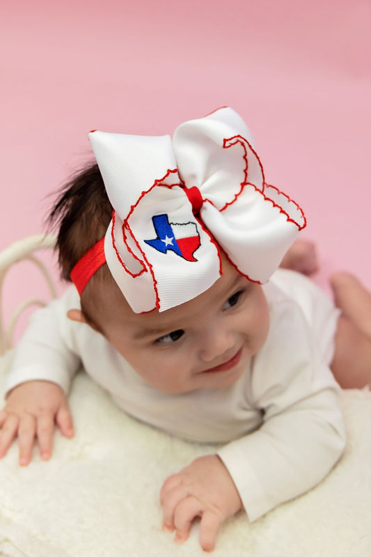 A baby wearing a white bow headband with a Texas flag motif in the center.
