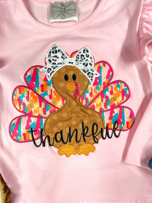 A light pink children's shirt featuring an embroidered turkey with colorful, patterned feathers in shades of pink, orange, blue, and yellow. The turkey has a white and black leopard-print bow on its head and the word 'thankful' stitched in black across its body. - Alternative view