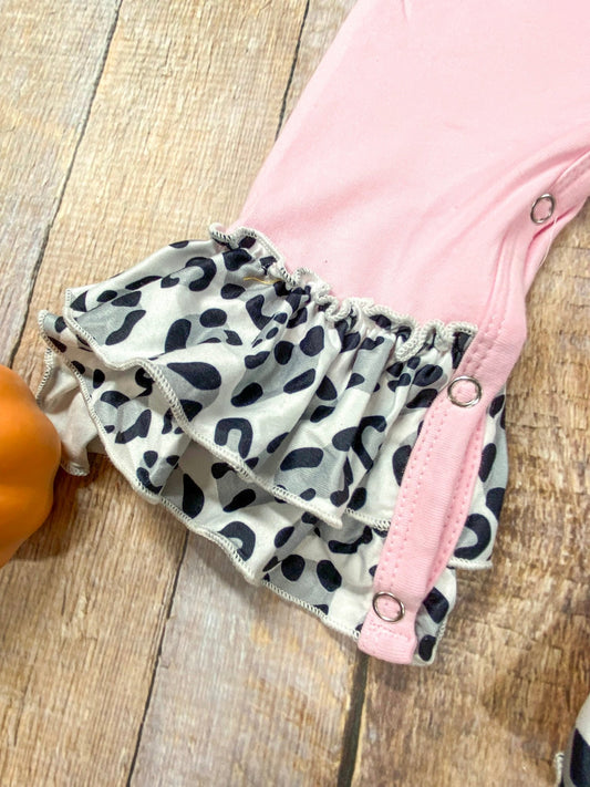 Close-up of a pink baby romper with snap buttons and layered ruffle leg cuffs featuring a black and white animal print pattern, displayed on a wooden surface next to a small orange pumpkin. - Alternative view