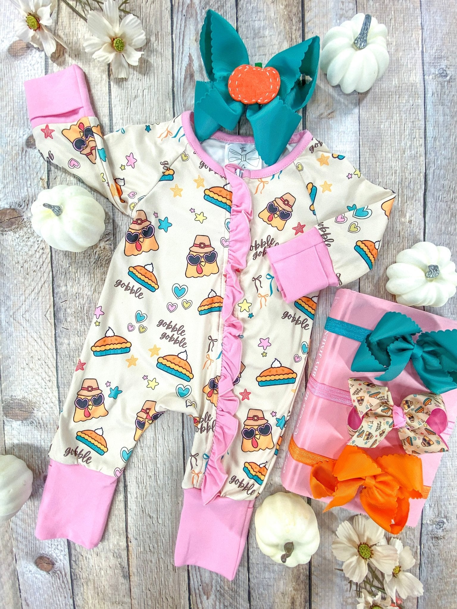 Flat lay of a baby girls’ long-sleeve romper designed for Thanksgiving. The cream-colored romper features playful cartoon turkeys wearing sunglasses and pilgrim hats, pumpkin pie slices, colorful stars, hearts, and the phrase ‘gobble gobble.’ It has pink cuffs, pink trim, and a pink ruffle running down the front zipper line. A teal bow with an orange felt pumpkin accent sits above the outfit, surrounded by white pumpkins, daisies, and pink gift boxes tied with vibrant bows