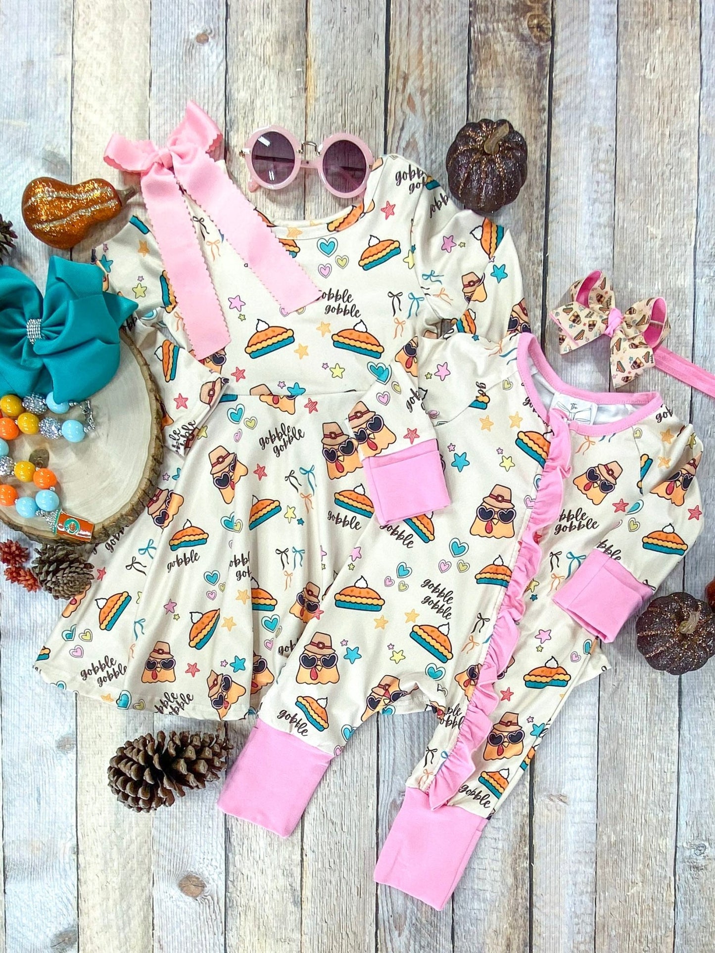 Flat lay of matching Thanksgiving outfits for siblings, featuring a baby romper and a girls’ dress made from the same cream-colored fabric with whimsical turkey and pumpkin pie prints, stars, hearts, and the words ‘gobble gobble.’ The romper has long sleeves with pink cuffs and ruffle trim, while the dress has long sleeves and a flared skirt. Styled together with coordinating bows and surrounded by autumn décor, pumpkins, and leaves