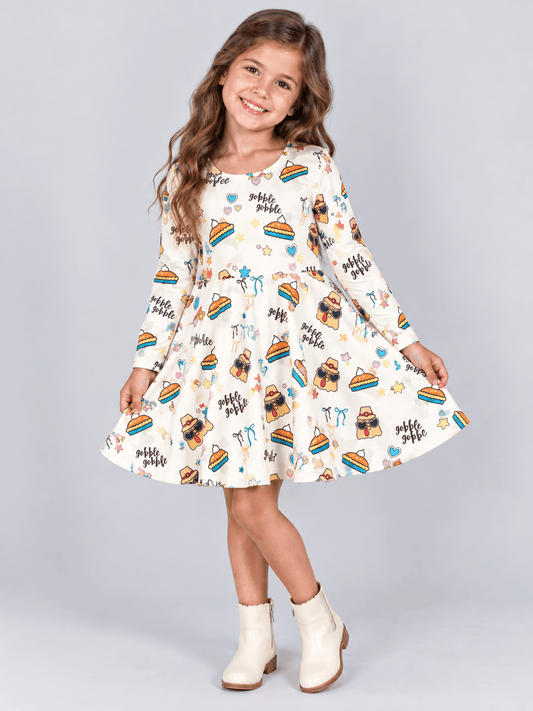 girls’ Thanksgiving dress made from cream-colored fabric with a fun holiday print of turkeys in sunglasses, pumpkin pie slices, colorful stars and hearts, and the words ‘gobble gobble'