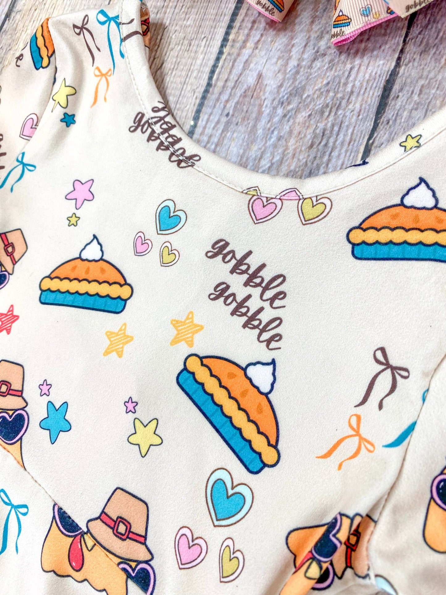 Close-up of the neckline on a girls’ Thanksgiving dress made from cream-colored fabric. The pattern features cheerful cartoon turkeys wearing sunglasses and pilgrim hats, slices of pumpkin pie topped with whipped cream, colorful stars and hearts, and the phrase ‘gobble gobble’ in dark brown script. The image highlights the smooth knit texture of the fabric and part of the rounded neckline with soft stitching visible in the background.