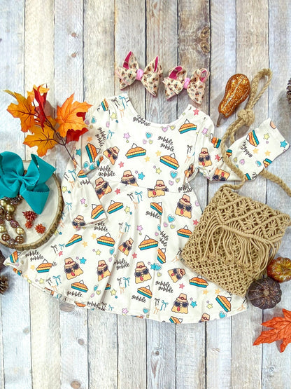 Flat lay of a girls’ Thanksgiving dress made from cream-colored fabric with a fun holiday print of turkeys in sunglasses, pumpkin pie slices, colorful stars and hearts, and the words ‘gobble gobble.’ The dress features long sleeves, a fitted bodice, and a full twirl skirt. Accessories include matching printed hair bows, a teal bow, a tan macramé purse, and fall decorations like leaves, pumpkins, and acorns arranged around the outfit.