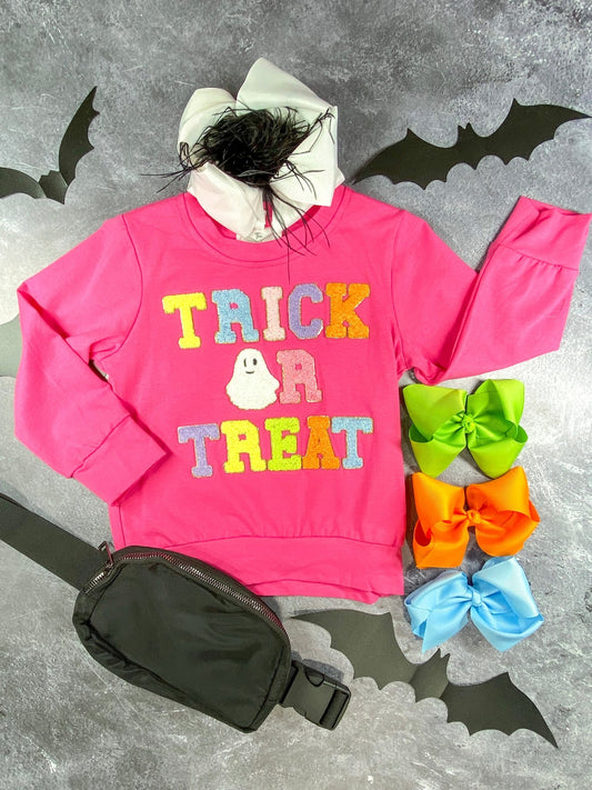 Flat lay of a bright pink children's sweatshirt with colorful, fuzzy letters spelling 'Trick or Treat' and a ghost illustration in place of the 'O'. The outfit is styled with a white bow featuring black feathers, a black waist bag, and three large hair bows in neon green, orange, and light blue (all sold separately)  - Alternative view